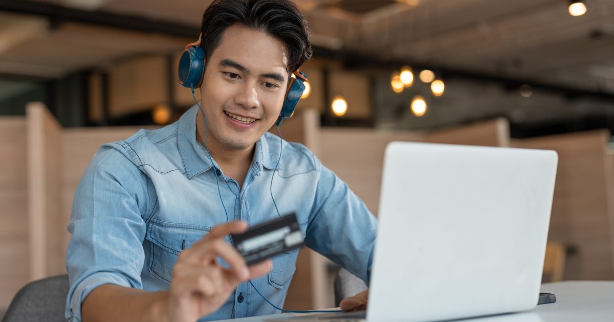 Young Asian male in blue shirt using credit card shopping online in laptop. Businessman making payment purchasing on internet.