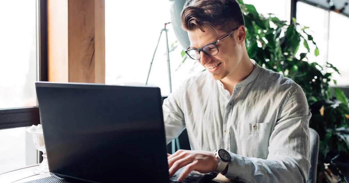 Attractive smiling businessman in eyeglasses working on laptop, typing, spending time at cafe. Dressed in white shirt.