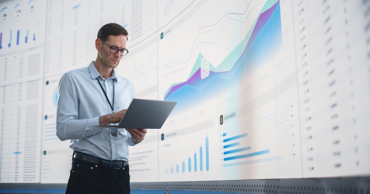 In The Office: Male Monitoring Specialist Holding Laptop Computer And Standing Next To Big Digital Screen With Graphs And Charts. Caucasian Man Analyzing Data, Performance Records, And Filing Report.