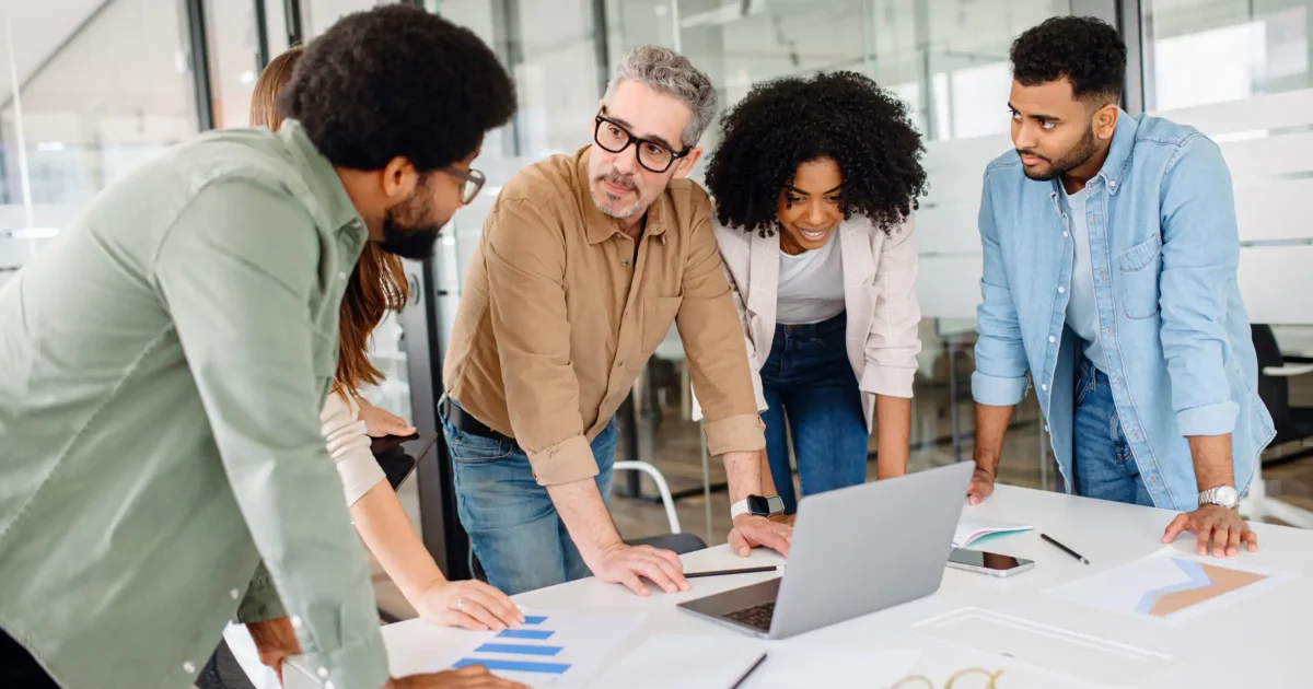 Team of professionals gathered around a laptop, analyzing data and discussing strategies in a modern office. A diverse group collaborating on a business project with documents, graphs, and technology